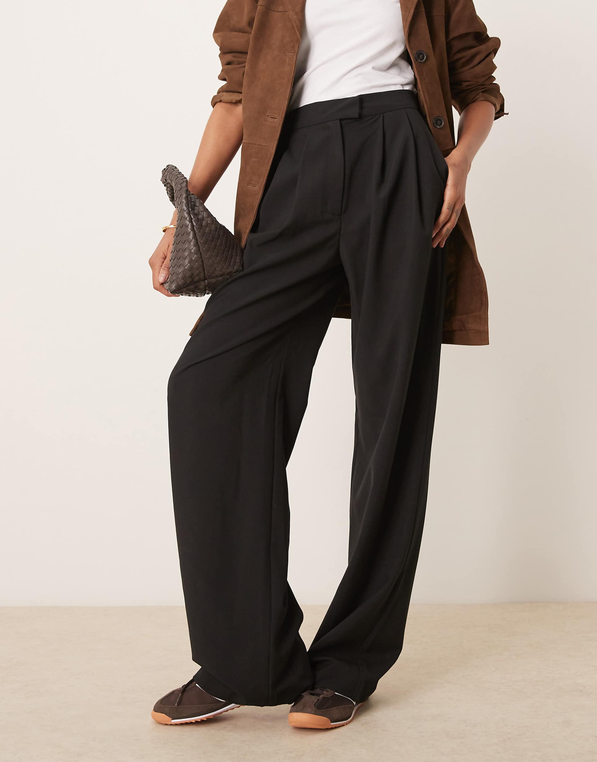 Person wearing black wide-leg pants with a brown coat and white shirt, holding a woven bag.