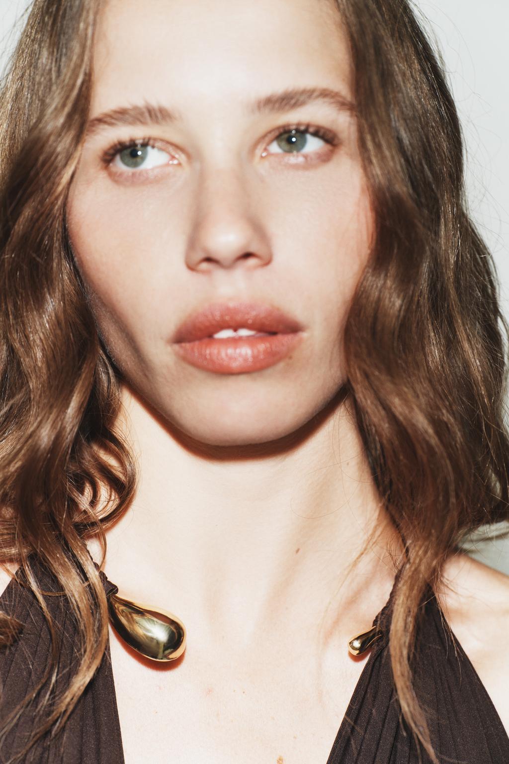 Close-up of a person with wavy hair wearing a necklace on a plain background